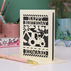 Personalised husband Birthday card with grey and cream design featuring a tractor and balloons, with 'Happy Birthday Edward' text, on a desk with stationery items.