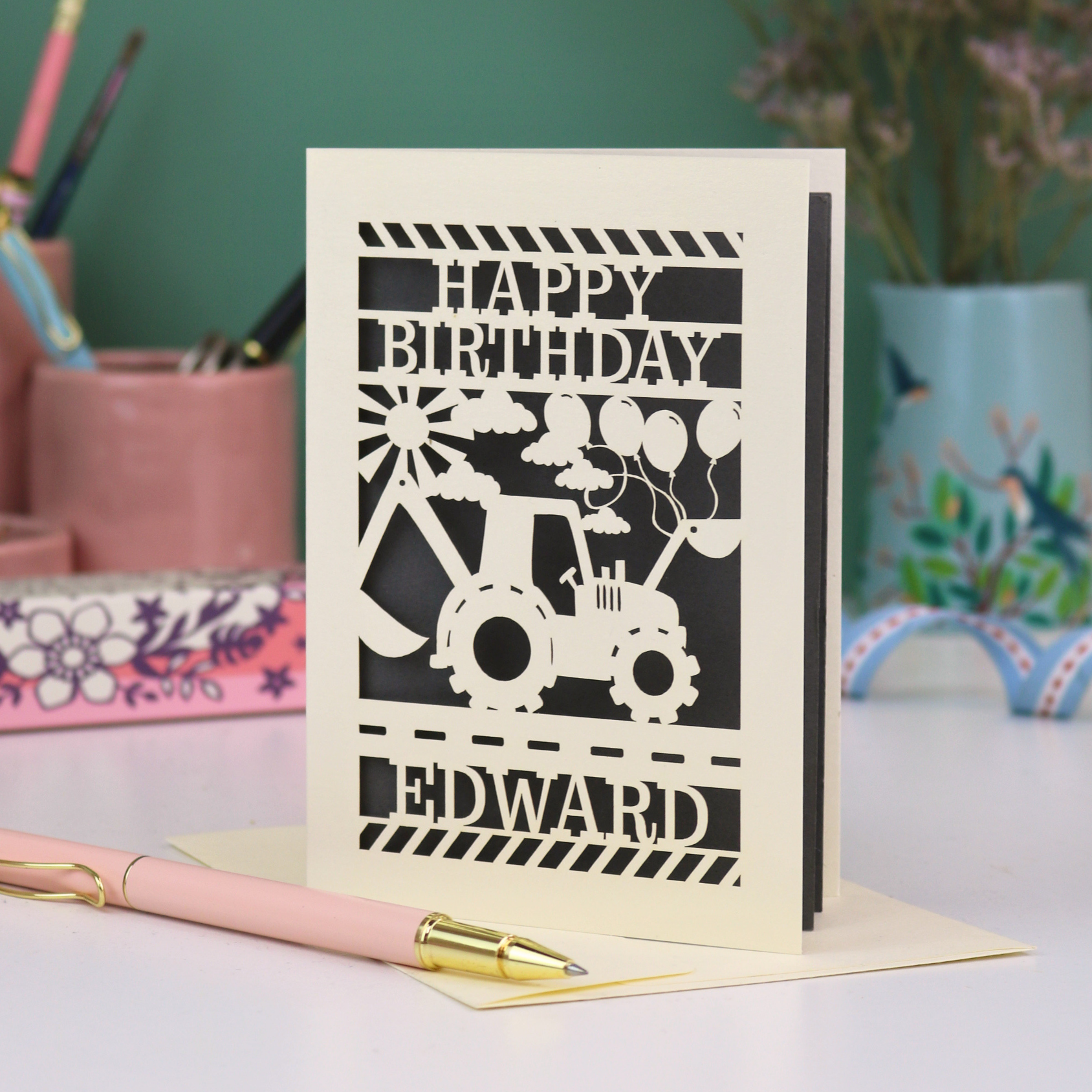 Personalised husband Birthday card with grey and cream design featuring a tractor and balloons, with 'Happy Birthday Edward' text, on a desk with stationery items.