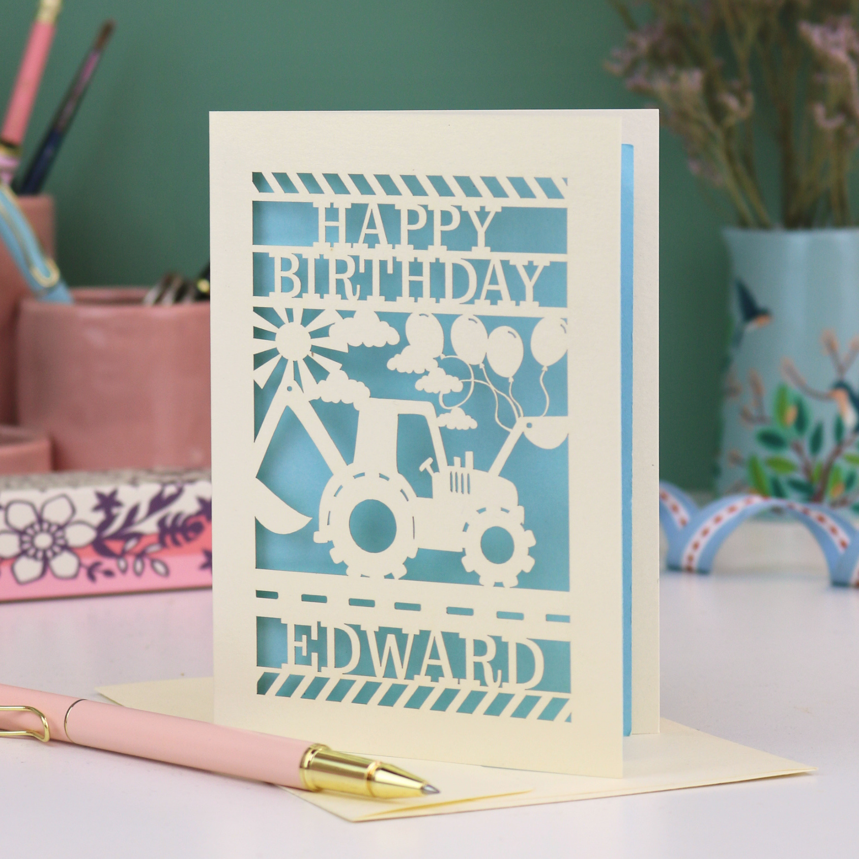 Personalised husband Birthday card with blue and cream design featuring a tractor and balloons, with 'Happy Birthday Edward' text, on a desk with stationery items.