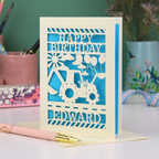 Personalised husband Birthday card with blue and cream design featuring a digger and balloons, with 'Happy Birthday Edward' text, on a desk with stationery items.