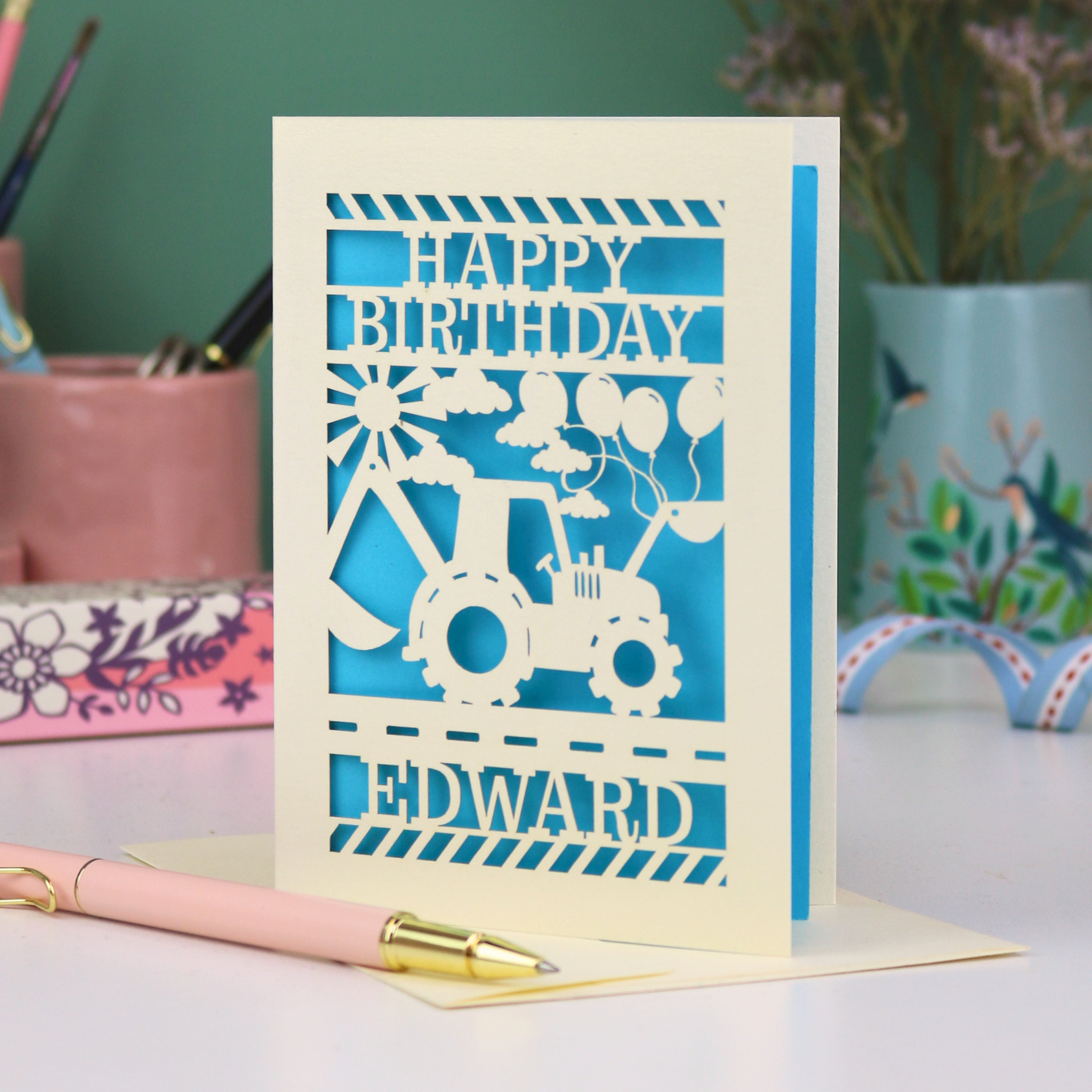 Personalised husband Birthday card with blue and cream design featuring a digger and balloons, with 'Happy Birthday Edward' text, on a desk with stationery items.
