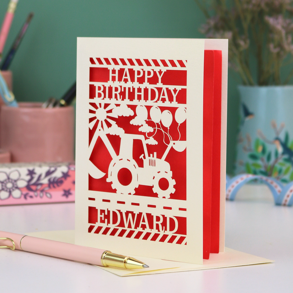 Personalised husband Birthday card with red and cream design featuring a tractor and balloons, with 'Happy Birthday Edward' text, on a desk with stationery items.