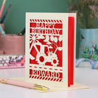 Personalised husband Birthday card with red and cream design featuring a tractor and balloons, with 'Happy Birthday Edward' text, on a desk with stationery items.