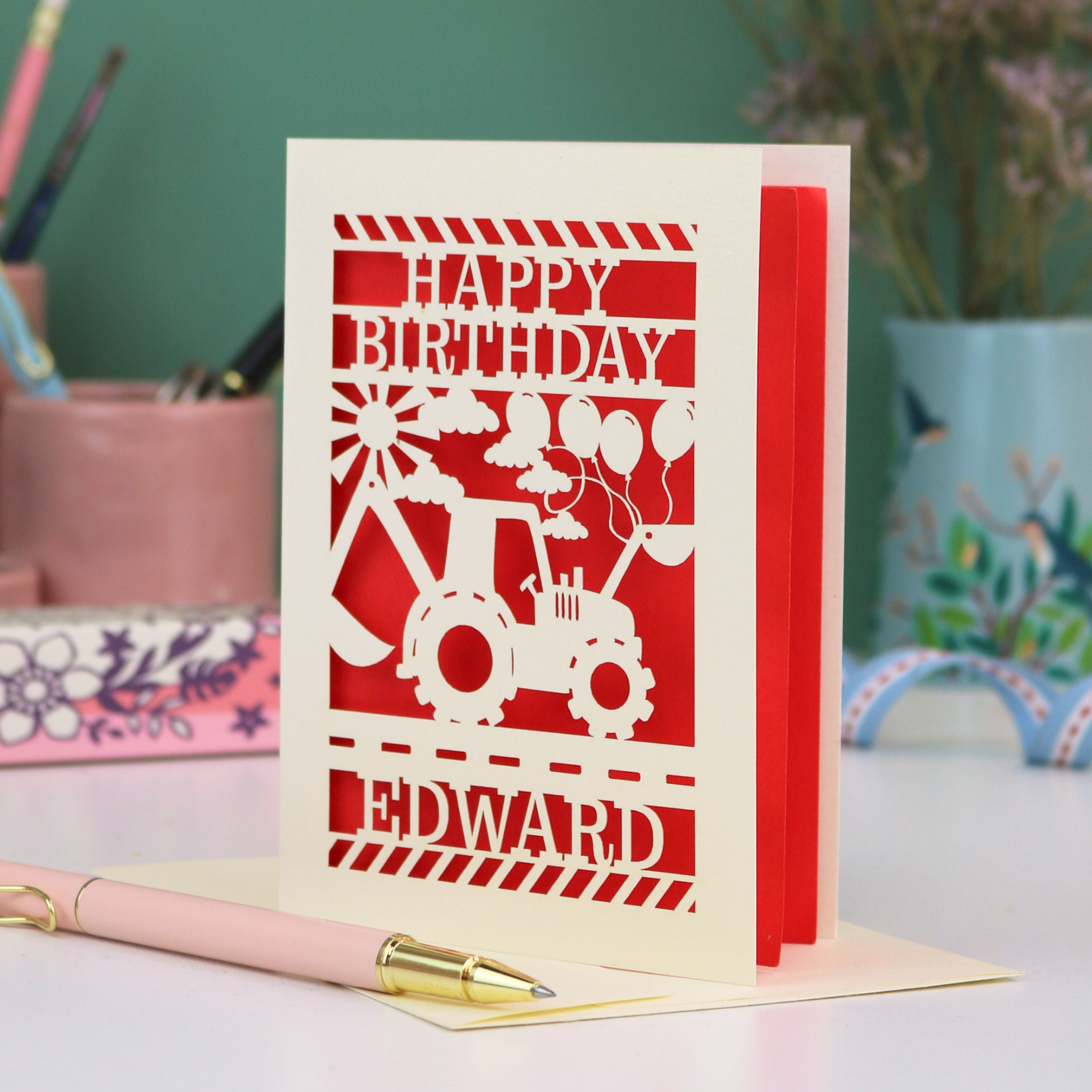 Personalised husband Birthday card with red and cream design featuring a tractor and balloons, with 'Happy Birthday Edward' text, on a desk with stationery items.