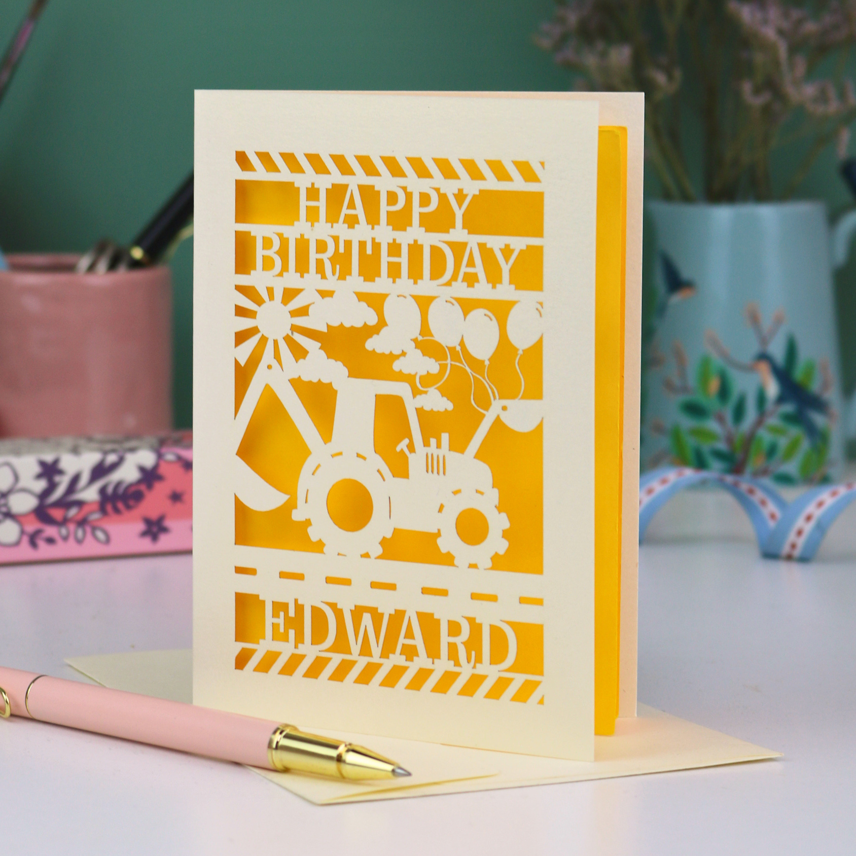 Personalised husband Birthday card with yellow and cream design featuring a tractor and balloons, with 'Happy Birthday Edward' text, on a desk with stationery items.