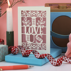 A laser cut "I love us" Valentine's card with ribbon, candle and pens on a white desk with pink background