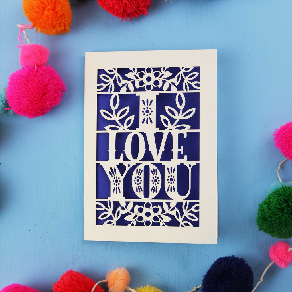 Paper cut Valentine's Card with 'I LOVE YOU' design surrounded by colourful pom-poms on a blue background