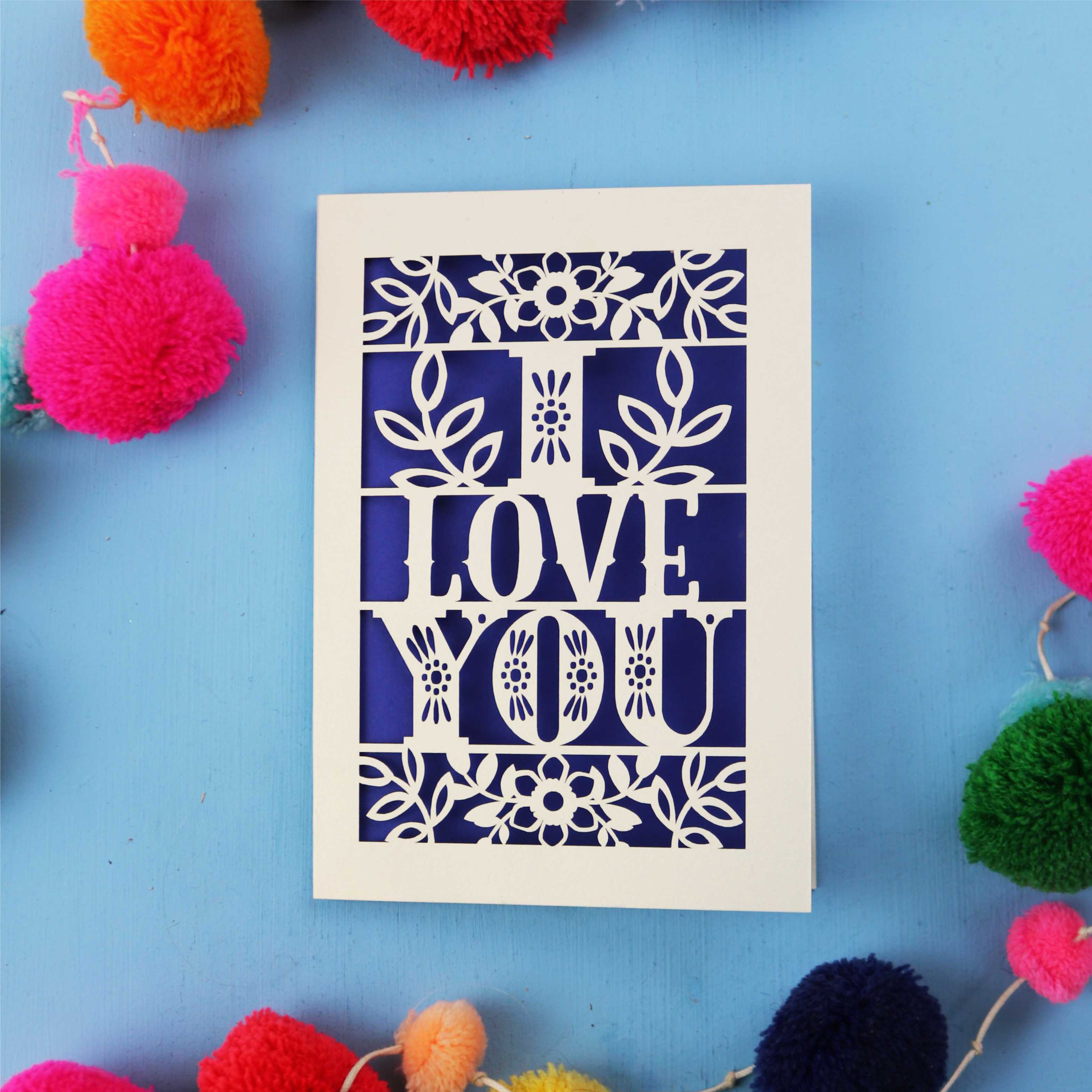 Paper cut Valentine's Card with 'I LOVE YOU' design surrounded by colourful pom-poms on a blue background