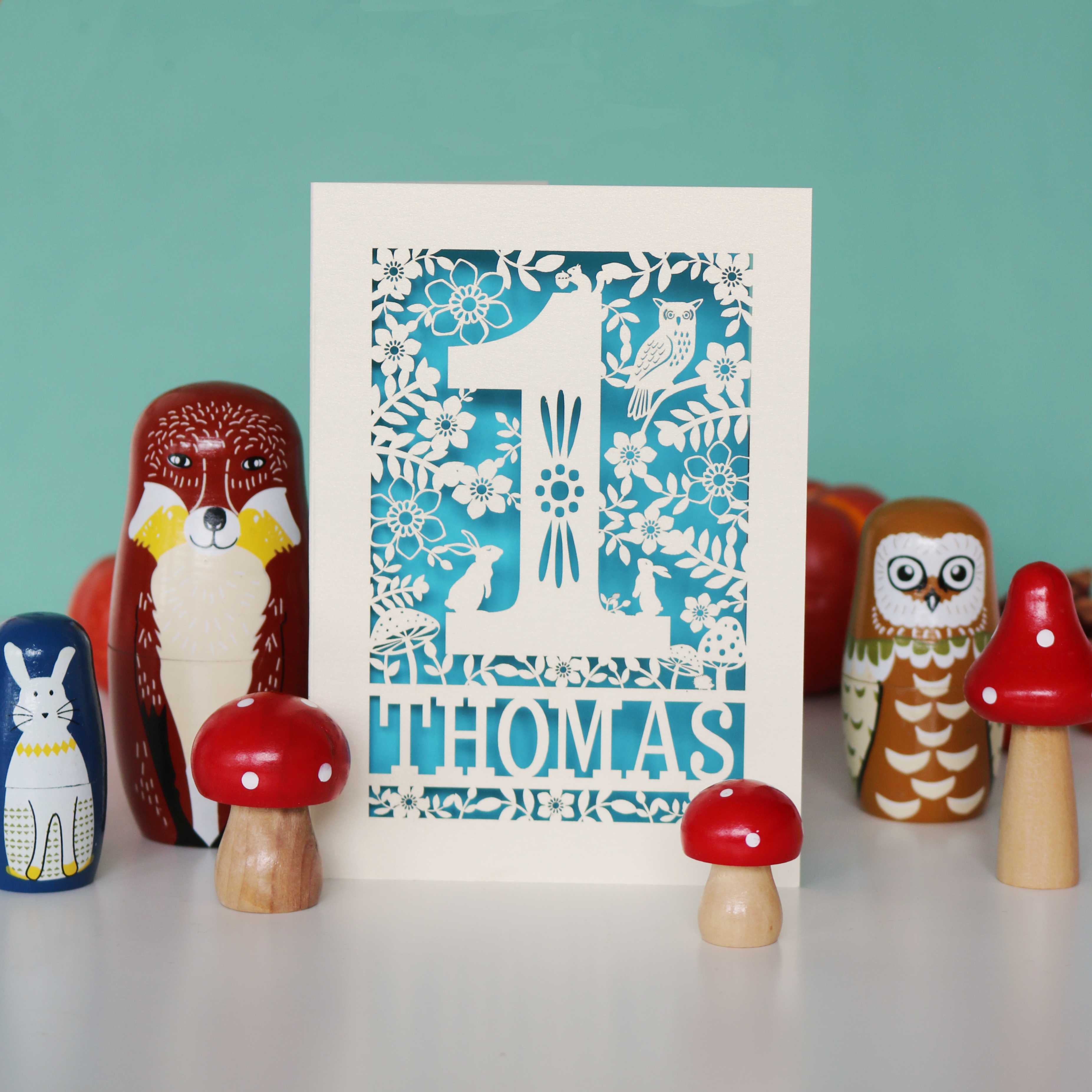 A personalised papercut 1st birthday card with a woodland animal theme, featuring cut-out shapes of an owl, rabbits, and a hedgehog, and the name 'THOMAS' in bold letters.