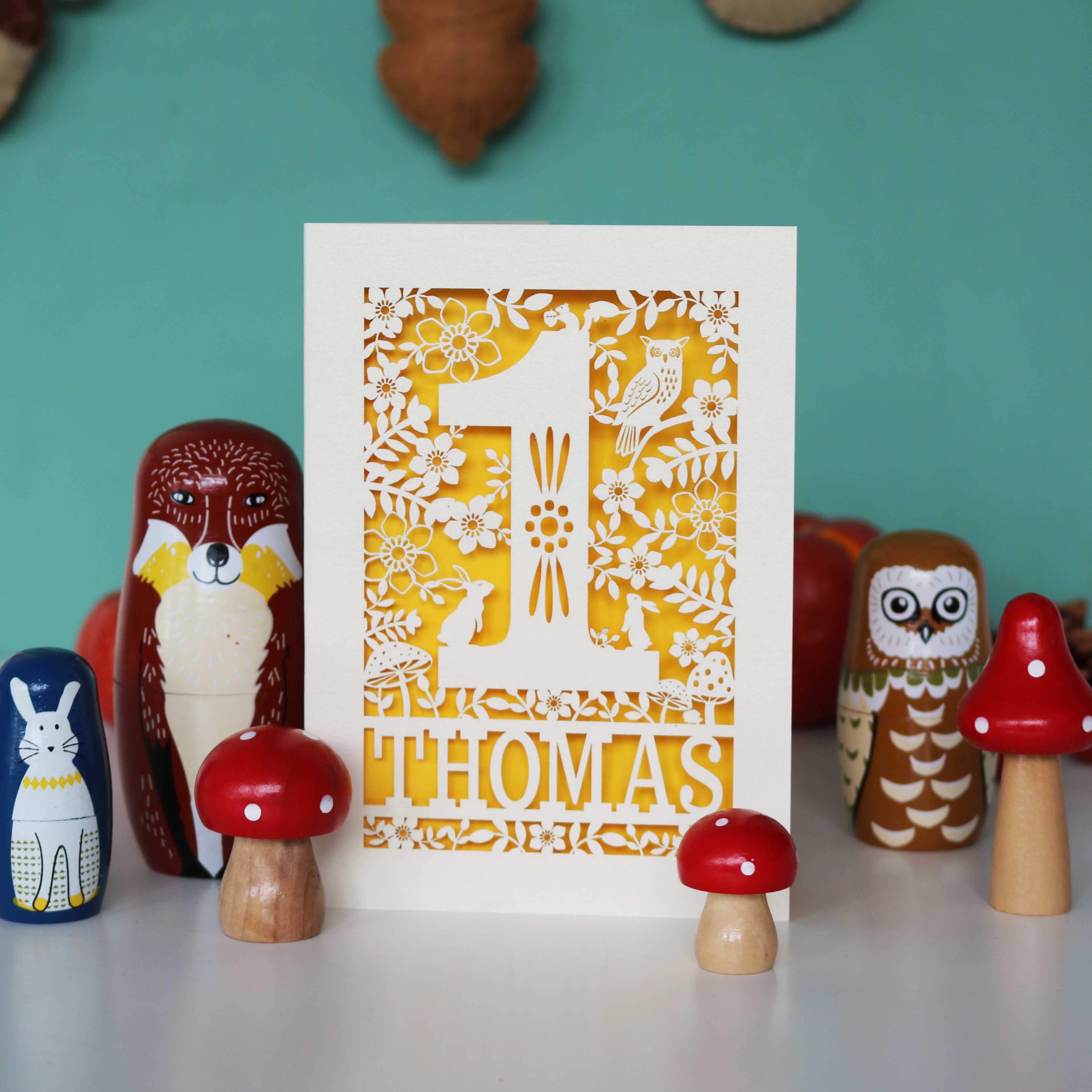 A personalised papercut 1st birthday card with a woodland animal theme, featuring cut-out shapes of an owl, rabbits, and a hedgehog, and the name 'THOMAS' in bold letters.