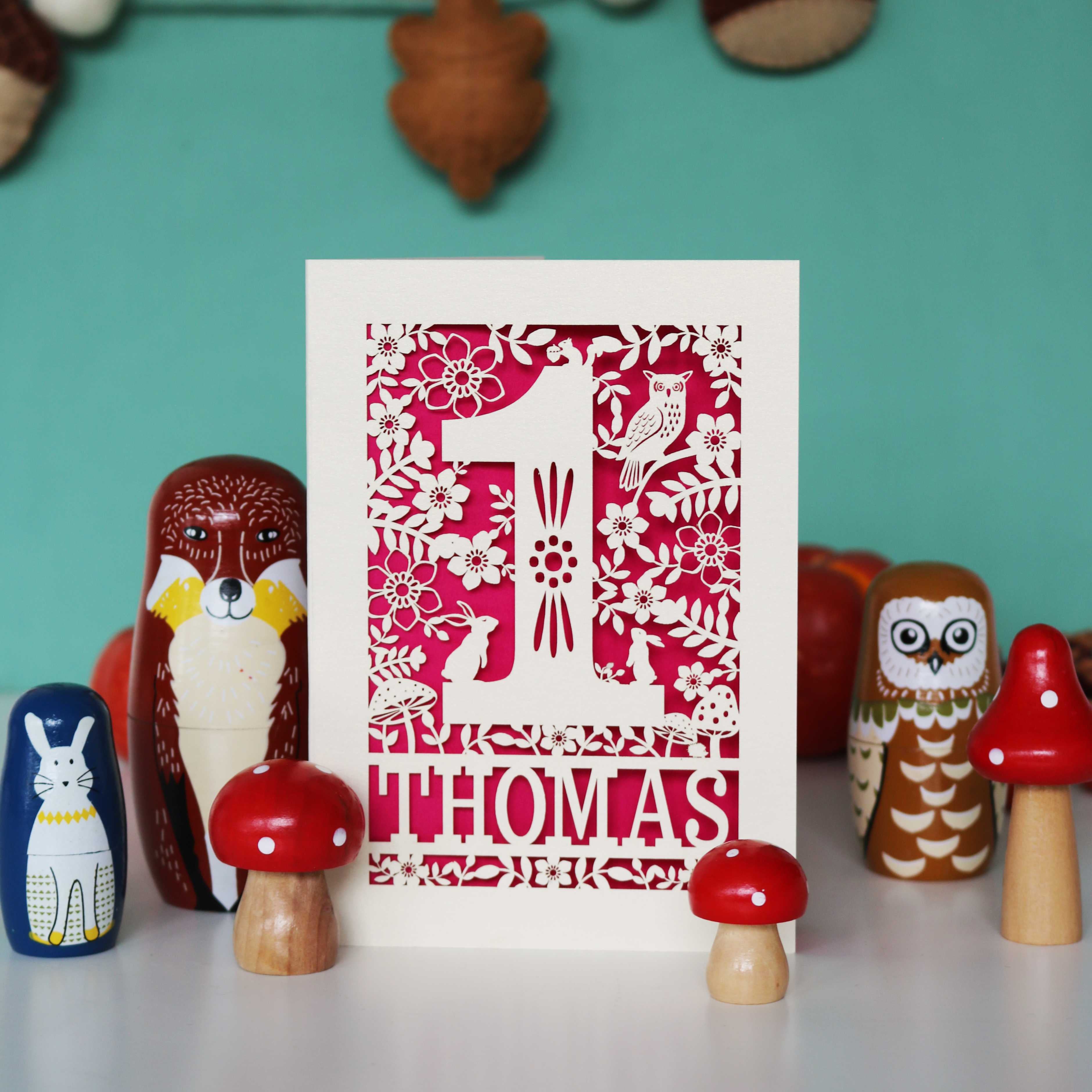 A personalised papercut 1st birthday card with a woodland animal theme, featuring cut-out shapes of an owl, rabbits, and a hedgehog, and the name 'THOMAS' in bold letters.