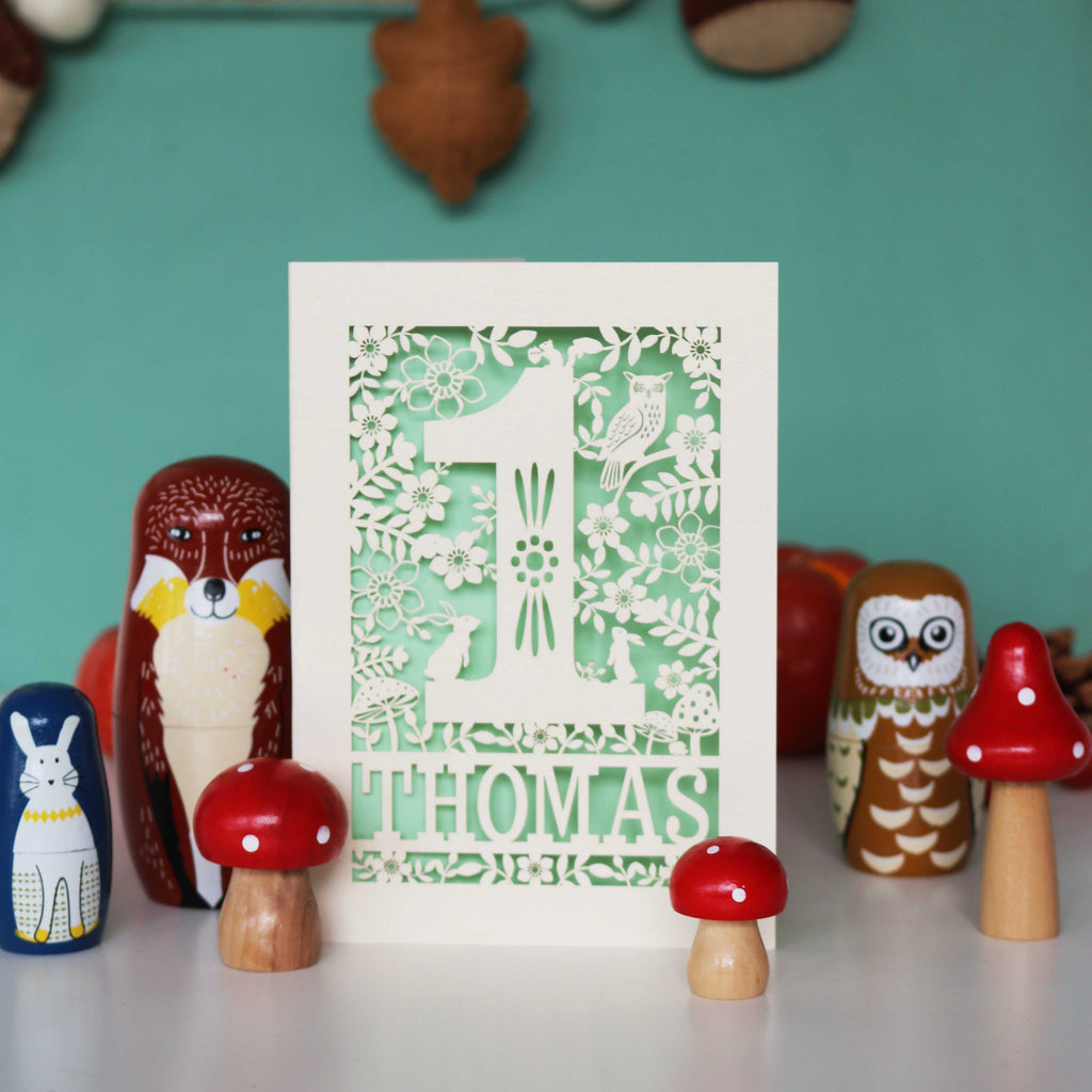 A personalised papercut 1st birthday card with a woodland animal theme, featuring cut-out shapes of an owl, rabbits, and a hedgehog, and the name 'THOMAS' in bold letters.
