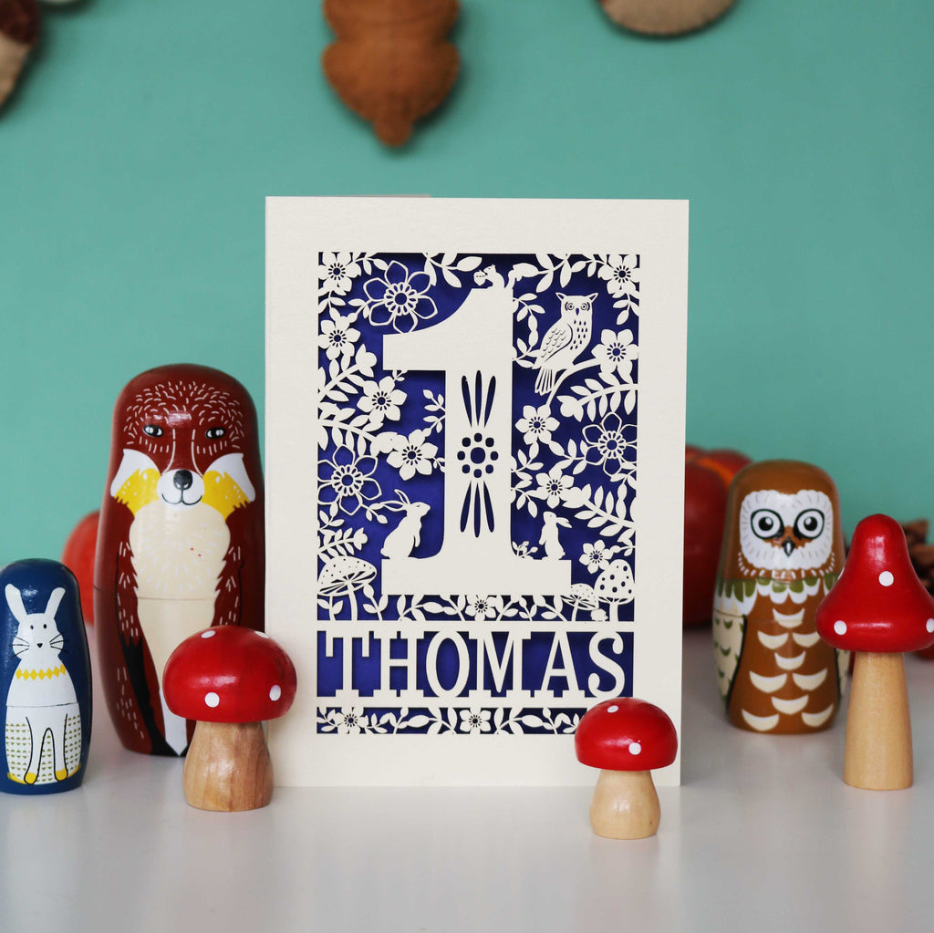 A personalised papercut 1st birthday card with a woodland animal theme, featuring cut-out shapes of an owl, rabbits, and a hedgehog, and the name 'THOMAS' in bold letters.