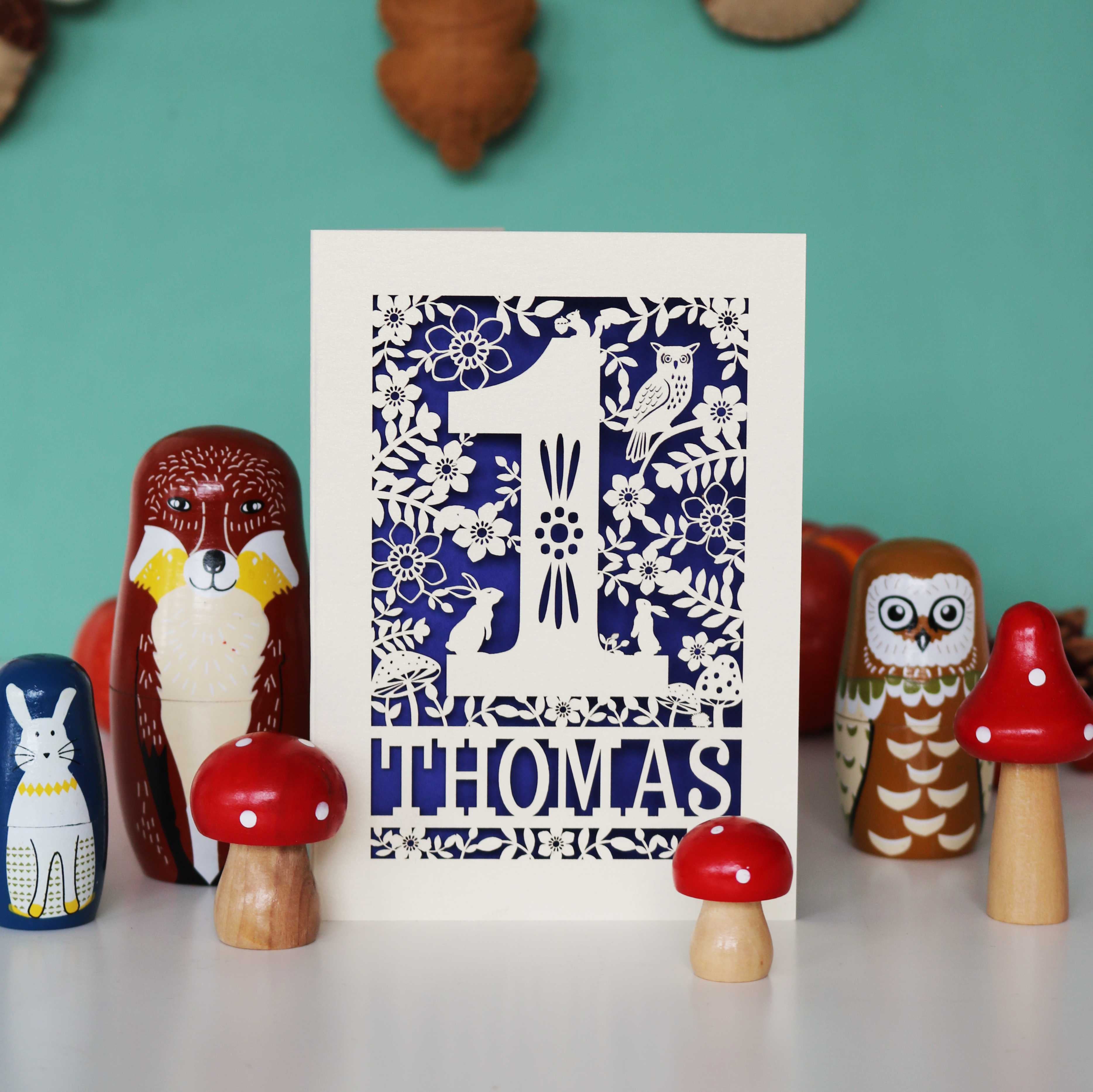 A personalised papercut 1st birthday card with a woodland animal theme, featuring cut-out shapes of an owl, rabbits, and a hedgehog, and the name 'THOMAS' in bold letters.