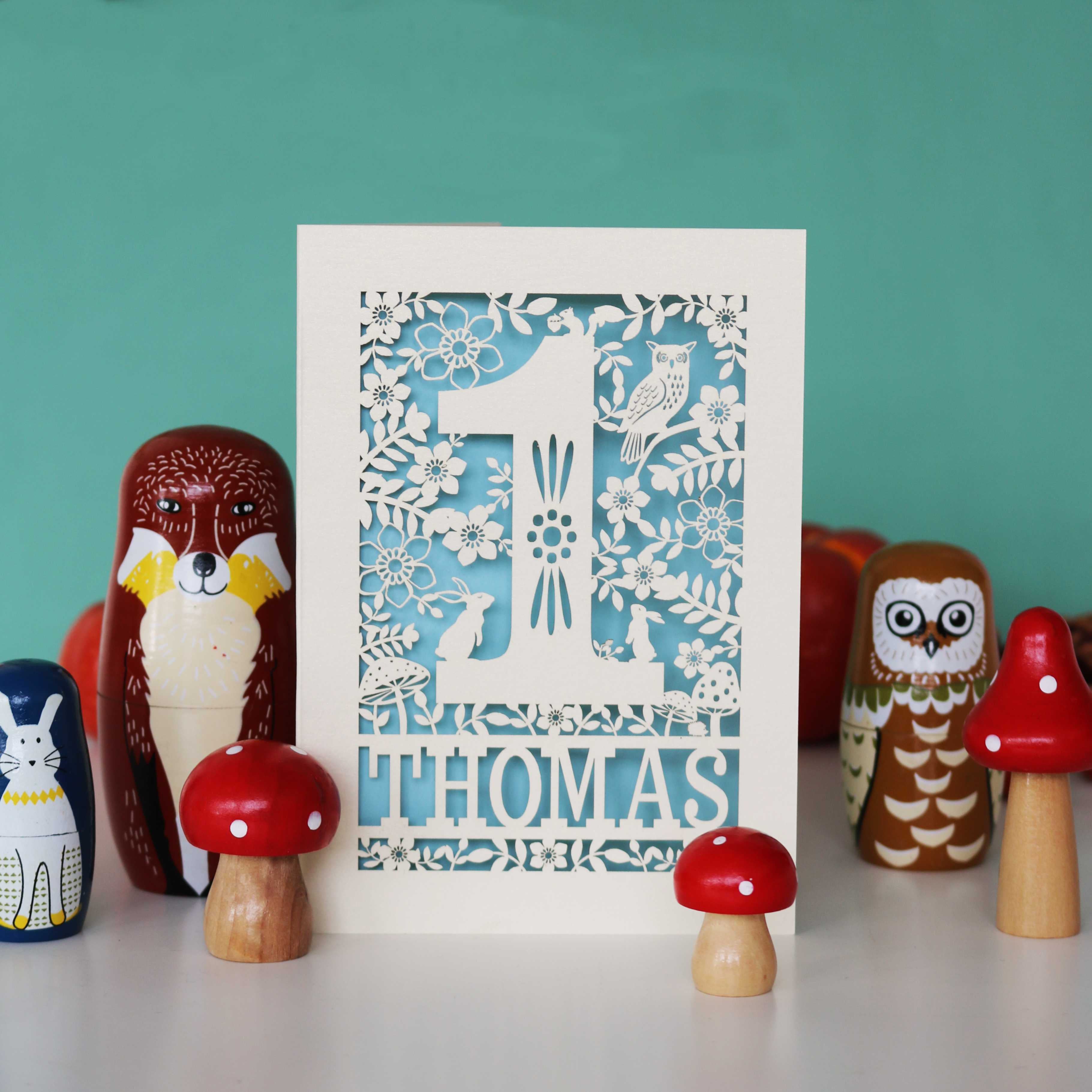 A personalised papercut 1st birthday card with a woodland animal theme, featuring cut-out shapes of an owl, rabbits, and a hedgehog, and the name 'THOMAS' in bold letters.