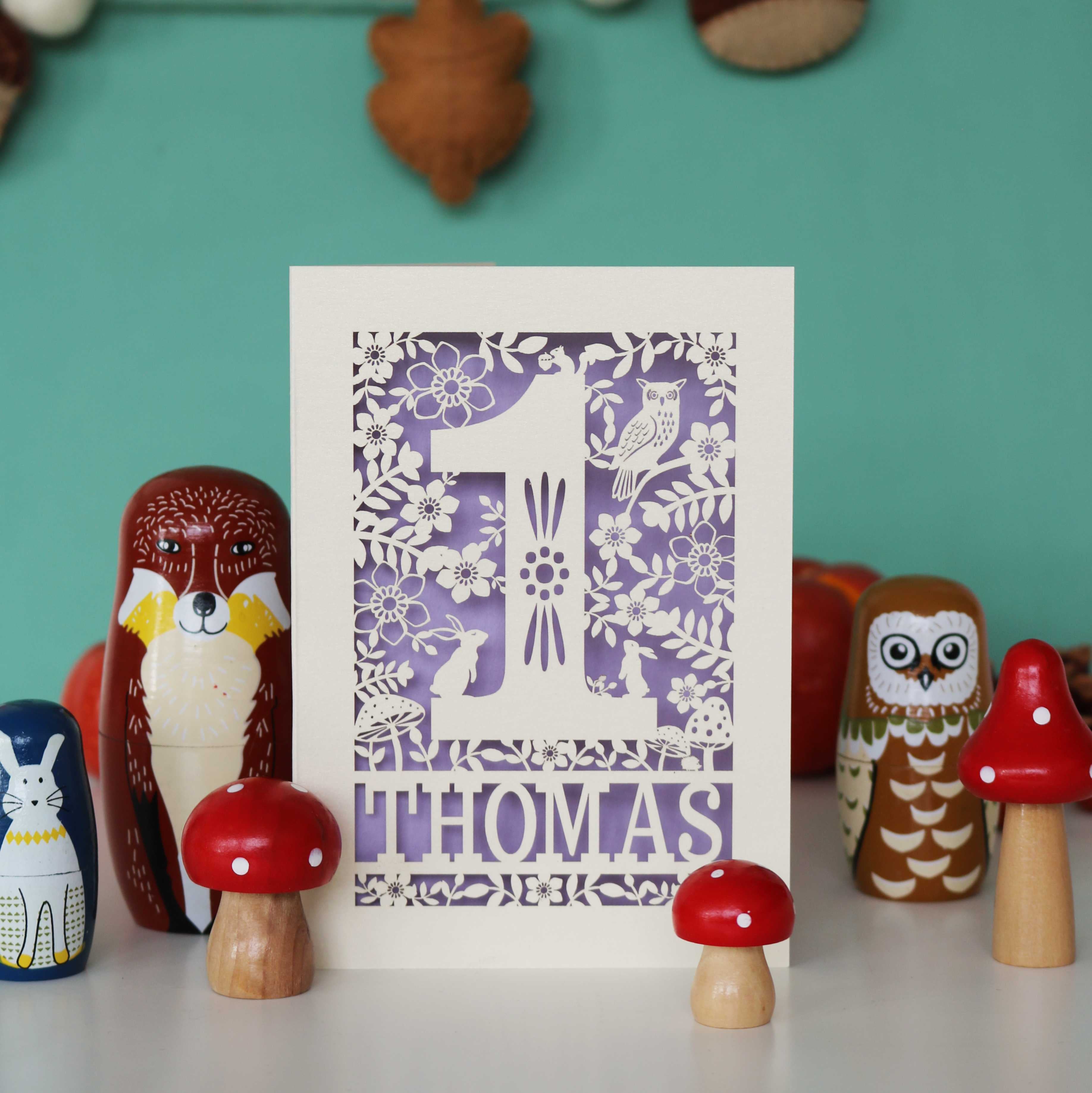 A personalised papercut 1st birthday card with a woodland animal theme, featuring cut-out shapes of an owl, rabbits, and a hedgehog, and the name 'THOMAS' in bold letters.