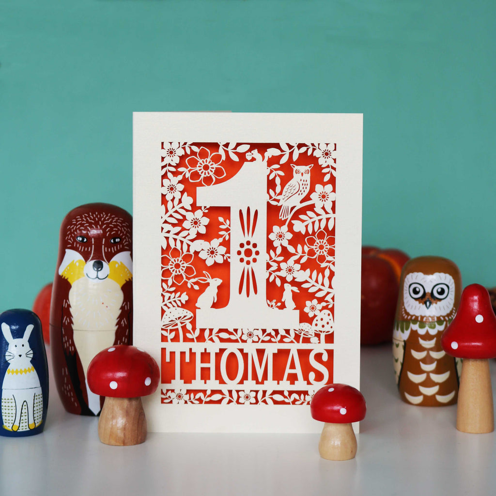 A personalised papercut 1st birthday card with a woodland animal theme, featuring cut-out shapes of an owl, rabbits, and a hedgehog, and the name 'THOMAS' in bold letters.