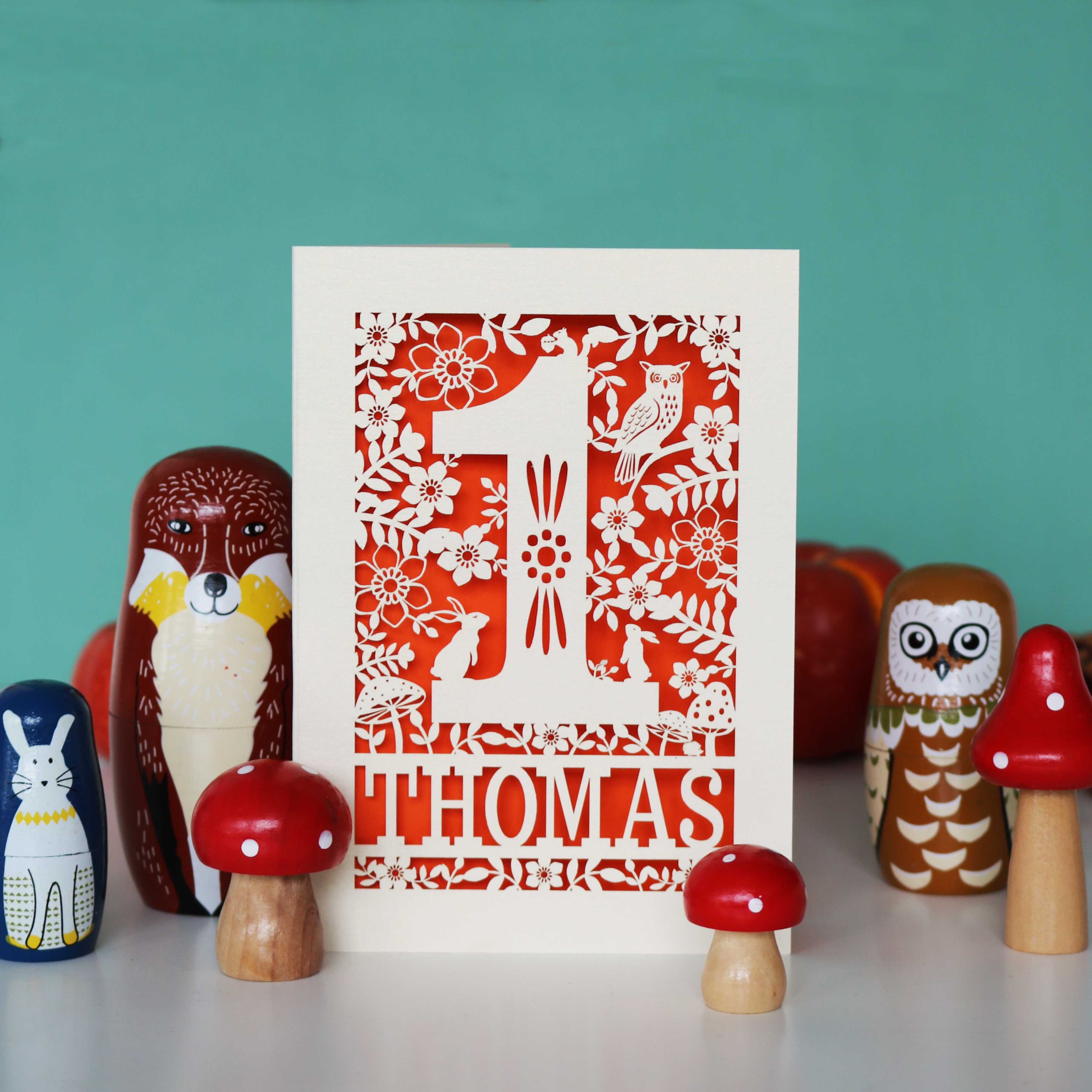 A personalised papercut 1st birthday card with a woodland animal theme, featuring cut-out shapes of an owl, rabbits, and a hedgehog, and the name 'THOMAS' in bold letters.