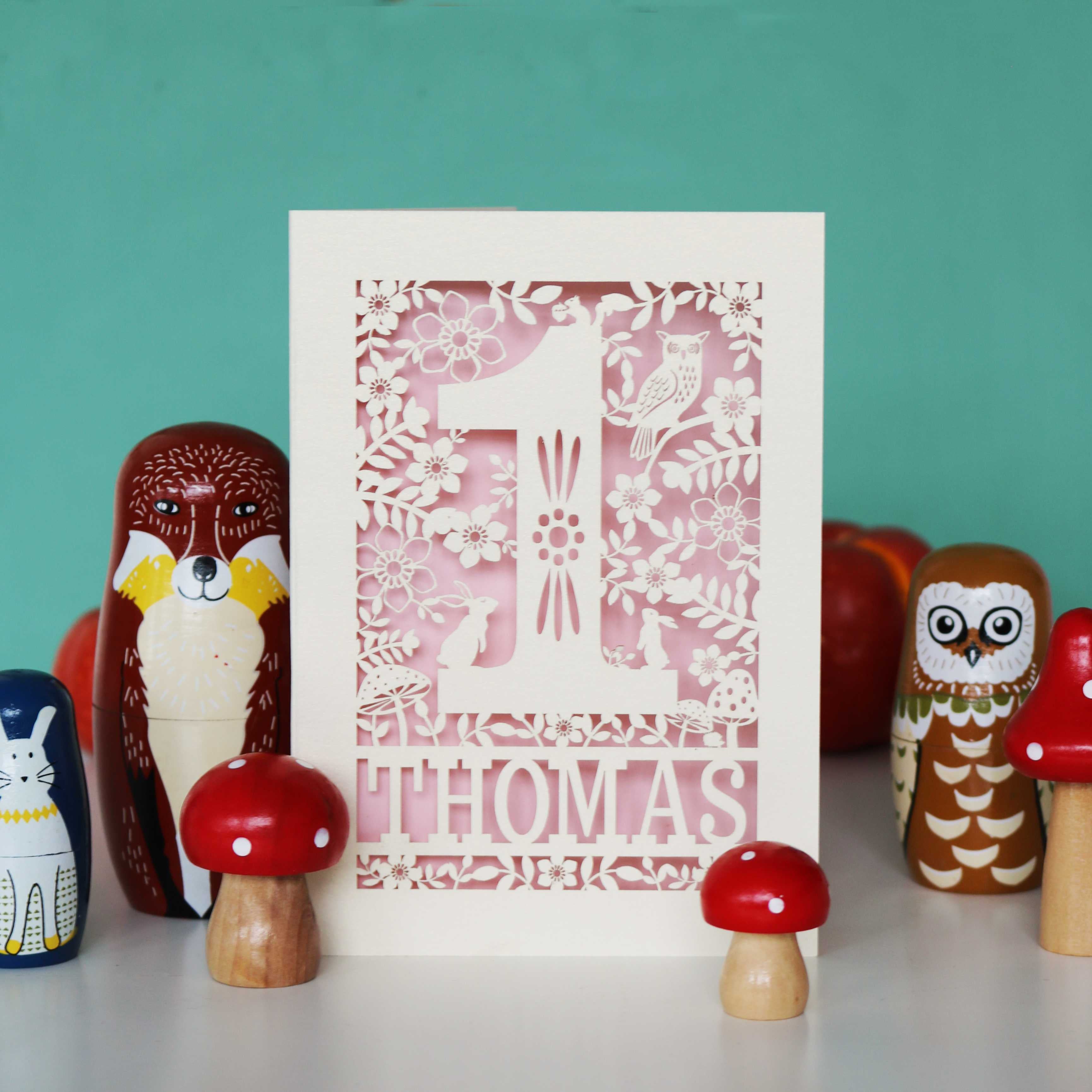 A personalised papercut 1st birthday card with a woodland animal theme, featuring cut-out shapes of an owl, rabbits, and a hedgehog, and the name 'THOMAS' in bold letters.