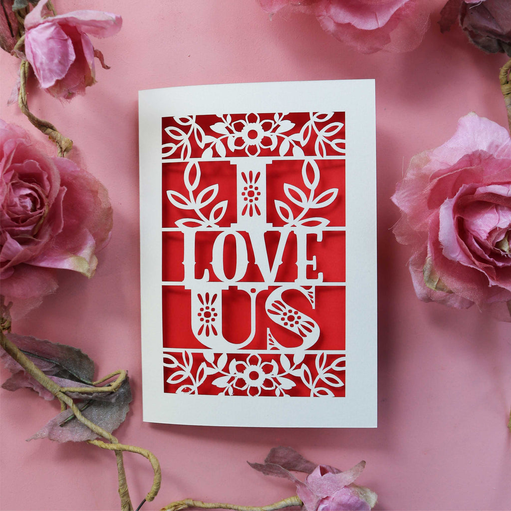 A papercut love card with the words 'I LOVE US' cut out in red, placed on a pink background with pink roses around it.