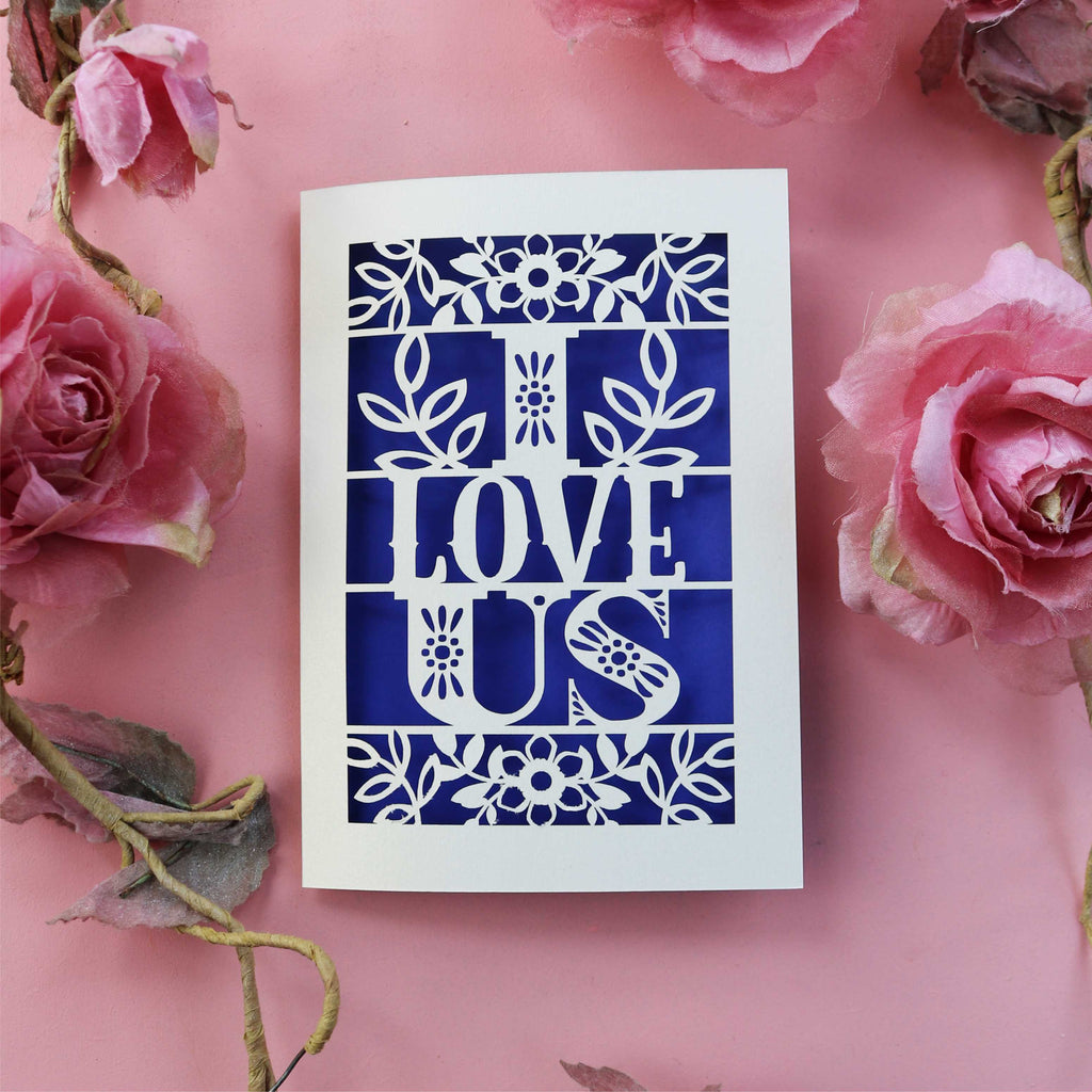 A papercut love Valentine's card with the words 'I LOVE US' cut out, placed on a pink background with pink roses around it.