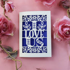A papercut love Valentine's card with the words 'I LOVE US' cut out, placed on a pink background with pink roses around it.