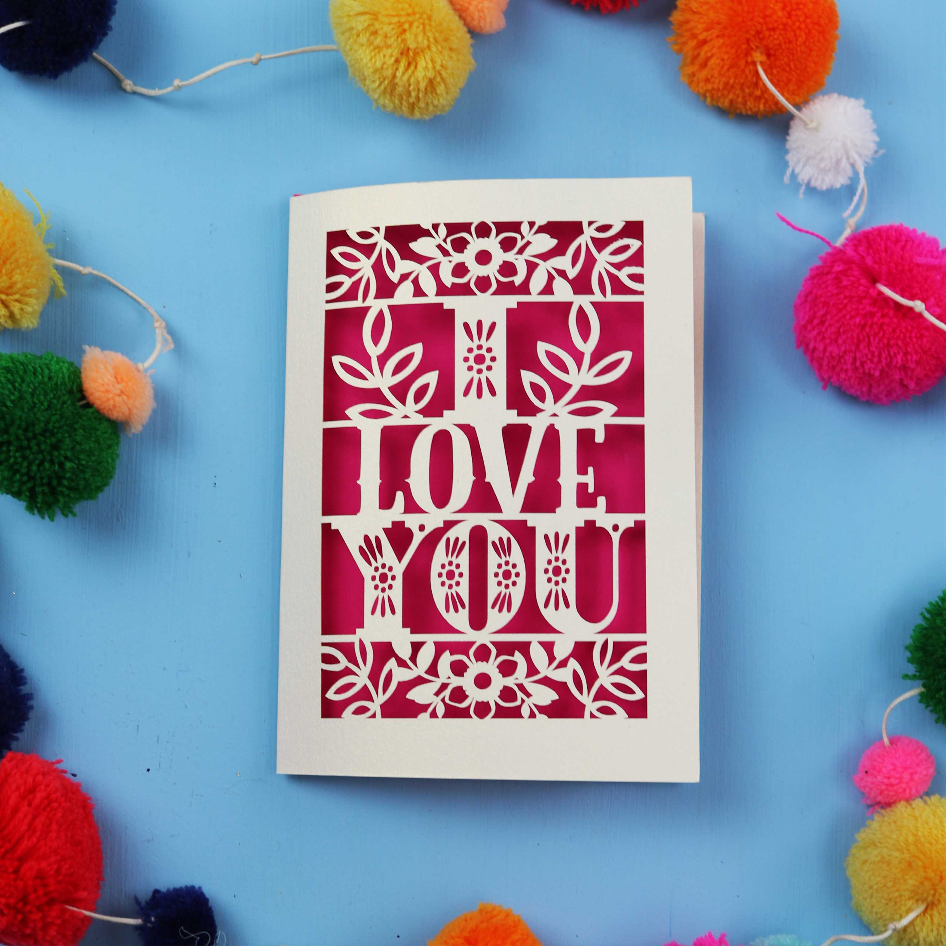 Laser cut Valentines Card with 'I LOVE YOU' design surrounded by colourful pom-poms on a blue background