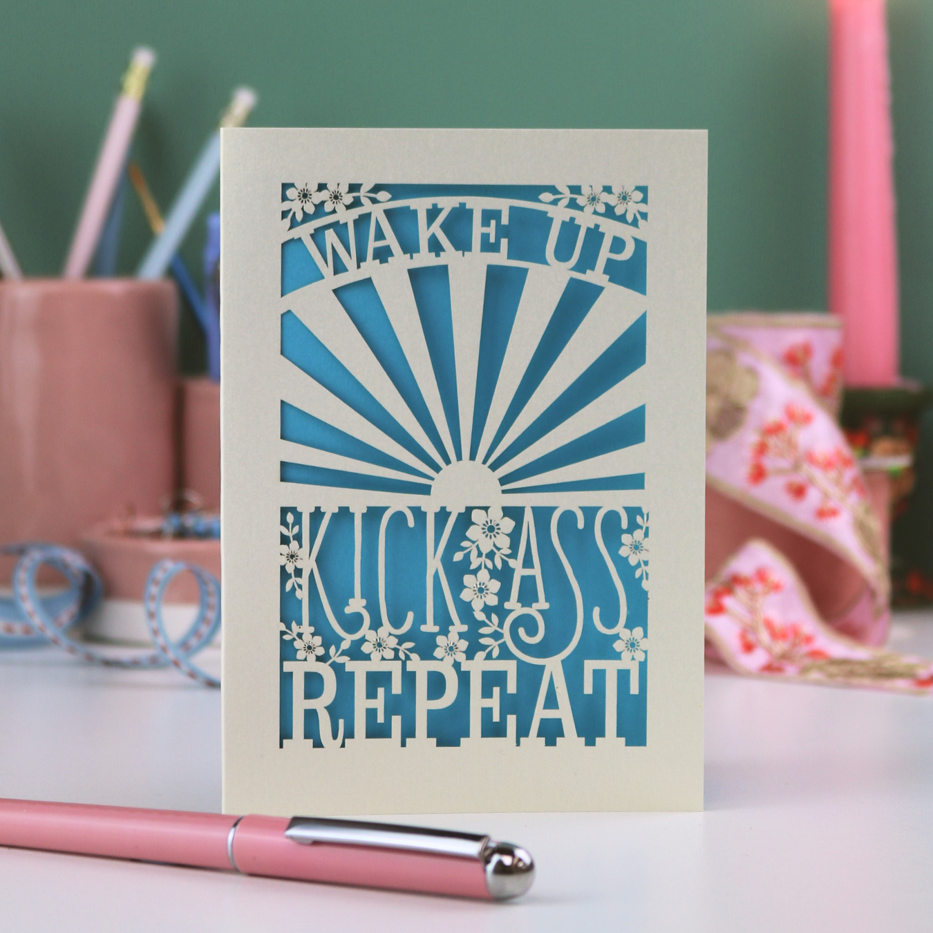A laser cut card with text "Wake up, kick ass, repeat" on a desk with stationery items