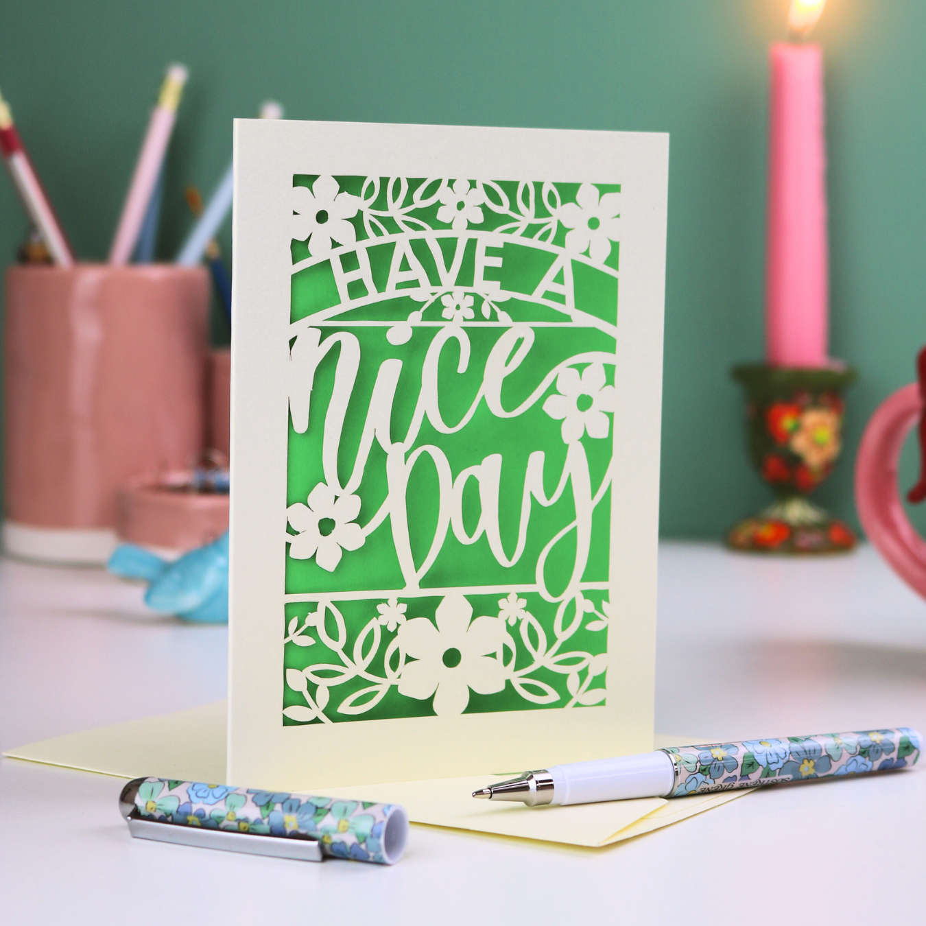 A laser cut card with text "Have a ice day", on a desk with stationery items
