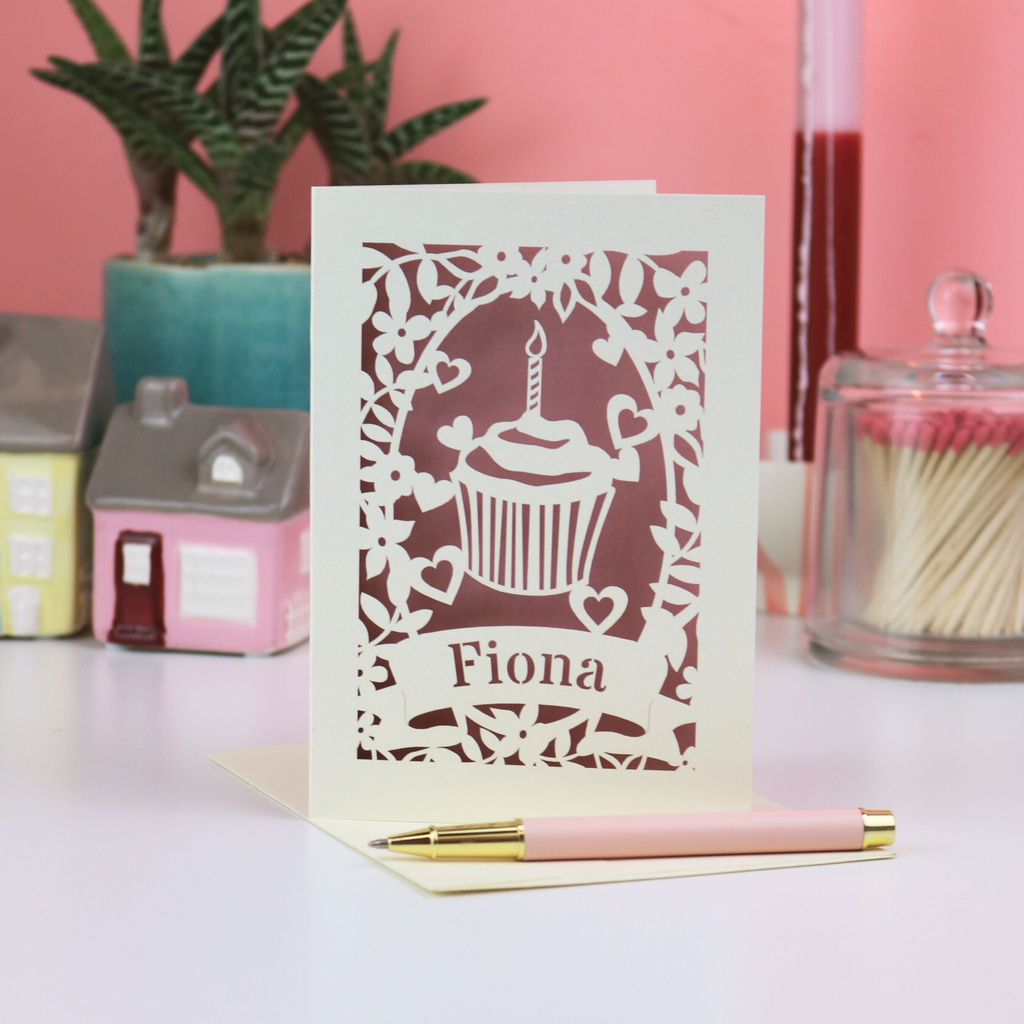 A laser cut cupcake birthday card with the name "Fiona", on white surface with pink background, plant, candle, matches and house decorative items