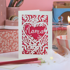 A cream and shocking pink laser cut card with cut out floral design and the word "Mama" in  heart shape. 