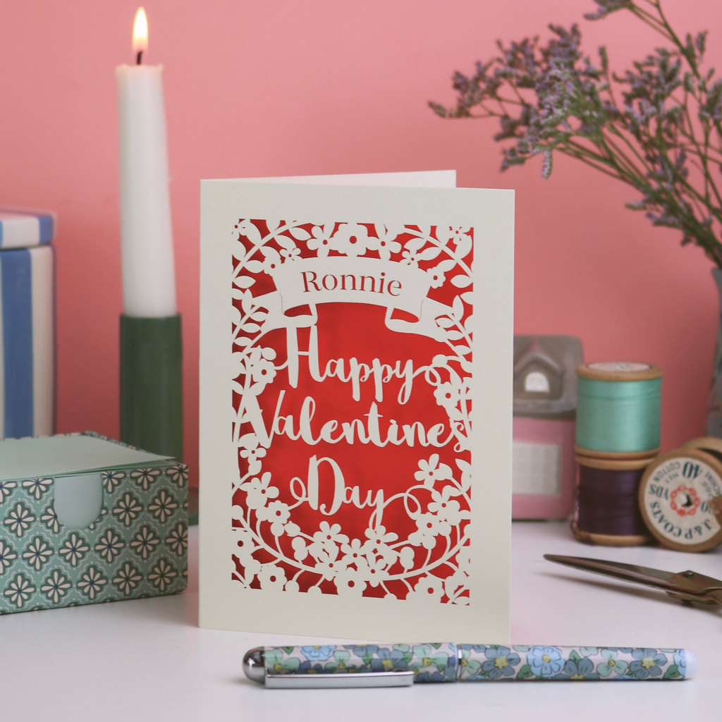 A personalised laser cut Valentine's Day card with decorative elements on a table with candles and flowers.