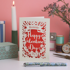 A personalised laser cut Valentine's Day card with decorative elements on a table with candles and flowers.