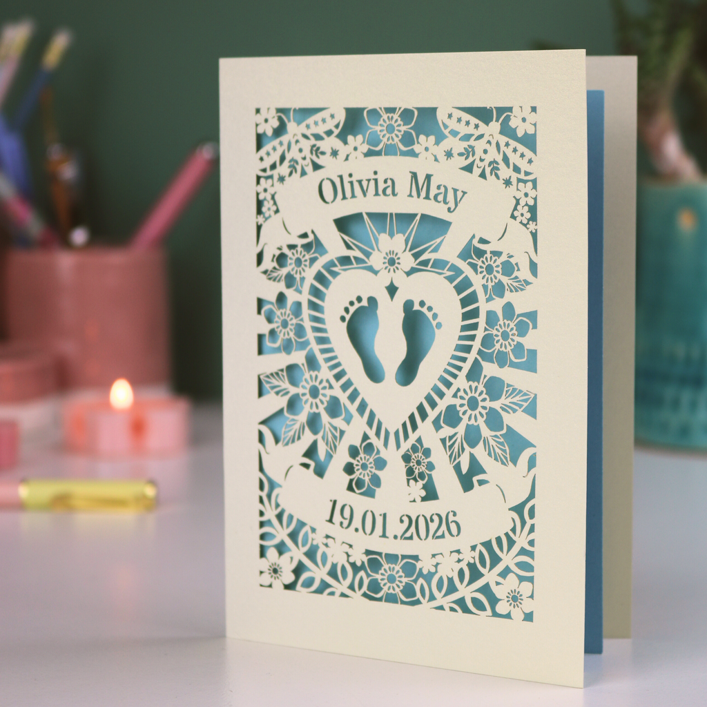 Baby card with laser-cut design, baby feet, and date on a table with stationery items.