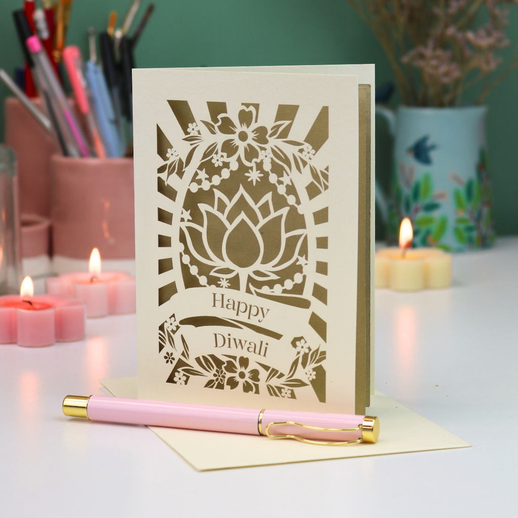 A decorative card with laser-cut floral and lotus designs and the words 'Happy Diwali' stands on a desk, with a pink pen resting in front. The scene includes lit candles, stationery in the background, and a green wall.