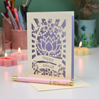 A cream-colored Diwali greeting card with a laser-cut lotus and floral design, standing upright on a desk with a pink pen resting in front. The card features cut out text 'Happy Diwali'