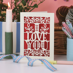 Card with 'I Love You' design on a table with candles and flowers in the background