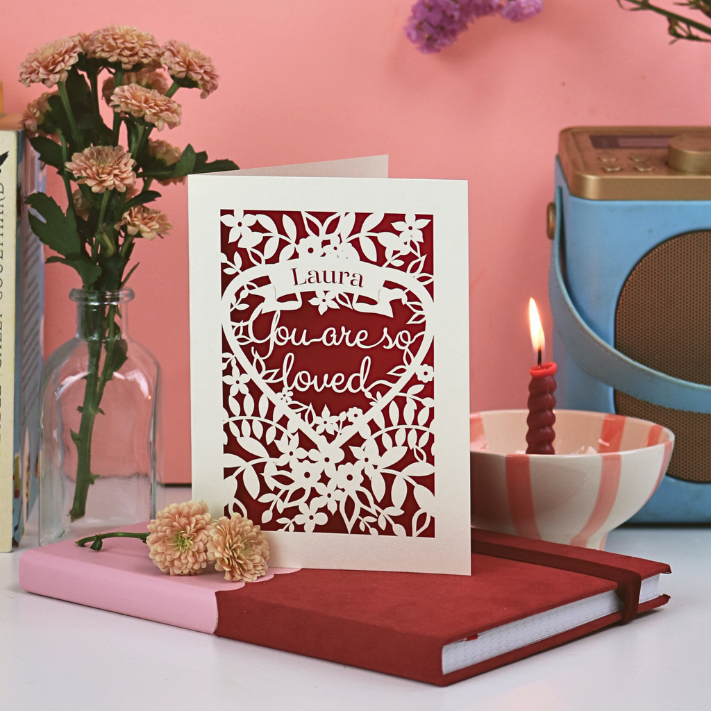 A laser cut Valentine's card with floral heart design and the words "Laura, you are so loved", on a pink background and desk setup