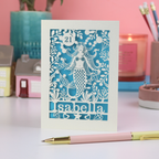 A laser cut mermaid birthday card, personalised with a name and age, on a white desk with stationery items, in front of a pink background