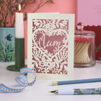Decorative laser cut Mother's Day card with floral design and 'Mum' text on a table with stationery items.