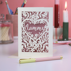 Laser-cut card with floral design and 'Mummy' text on a pink background with stationery items.