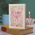 A laser cut card with floral design and cut out heart shape with "Nan" inside in a script font, standing on a yellow book with green background