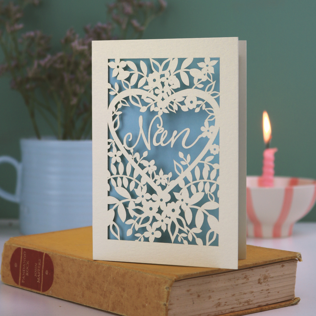 A laser cut card with floral design and cut out heart shape with "Nan" inside in a script font, standing on a yellow book with green background
