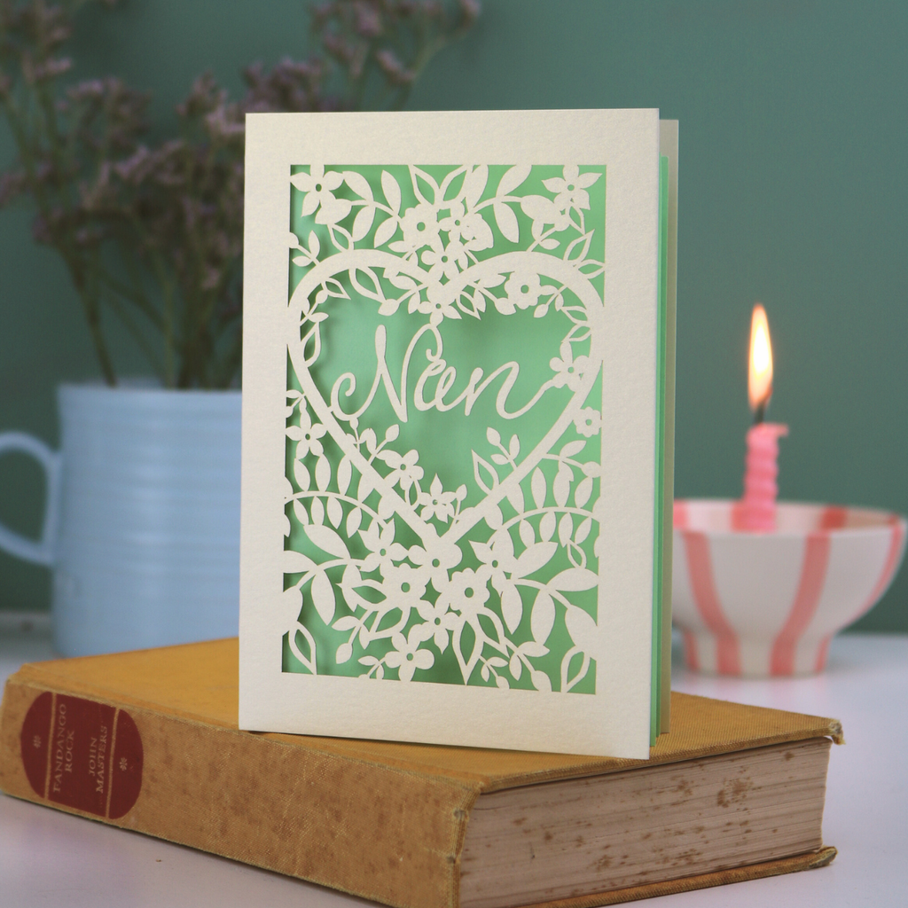 A laser cut Grandma card with floral design and cut out heart shape with "Nan" inside in a script font, standing on a yellow book with green background