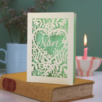 A laser cut Grandma card with floral design and cut out heart shape with "Nan" inside in a script font, standing on a yellow book with green background