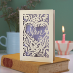 A laser cut Nan card with floral design and cut out heart shape with "Nan" inside in a script font, standing on a yellow book with green background