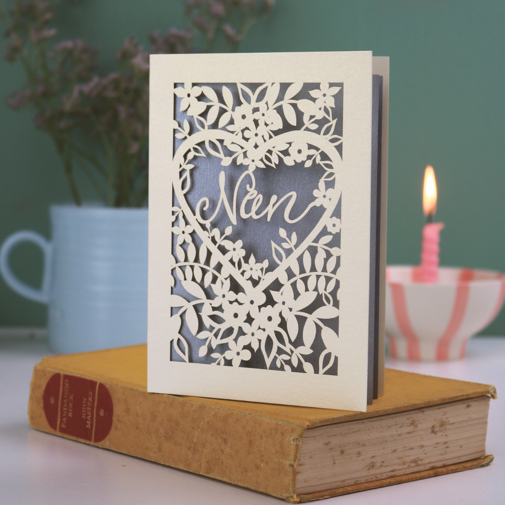 A laser cut greetings card with floral design and cut out heart shape with "Nan" inside in a script font, standing on a yellow book with green background