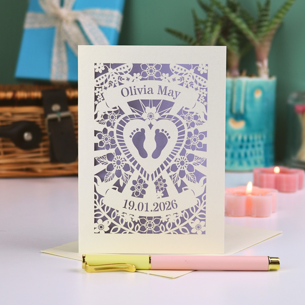 An intricate laser cut new baby card personalised with name and date, on a white desk with pen, plant and candles. 
