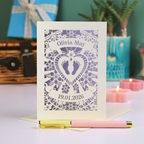 An intricate laser cut new baby card personalised with name and date, on a white desk with pen, plant and candles. 