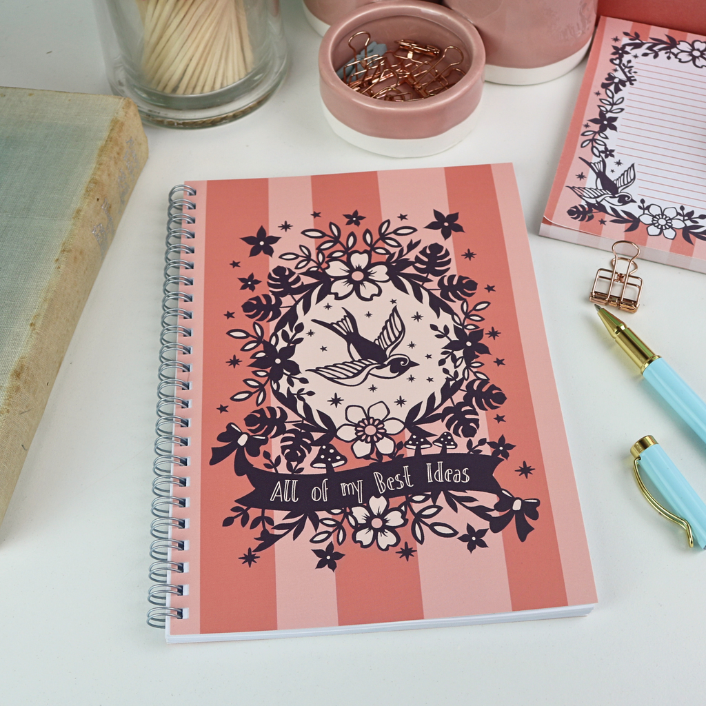 A wire bound A5 notebook with pink striped cover and purple floral illustrations, and the text "All of my Best Ideas", on a white desk with stationery items